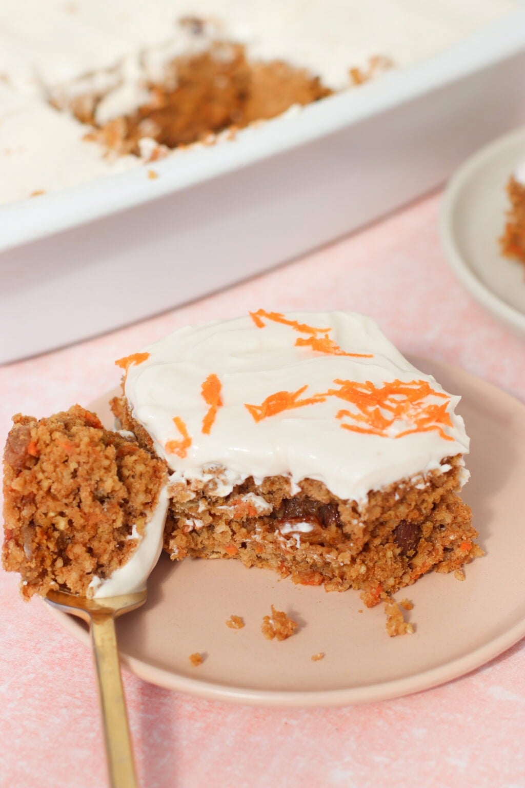 A slice of carrot cake with cream cheese frosting and grated carrot on top sits on a pink plate. A fork with a bite of cake rests next to it. The cake pan with more cake is visible in the background.