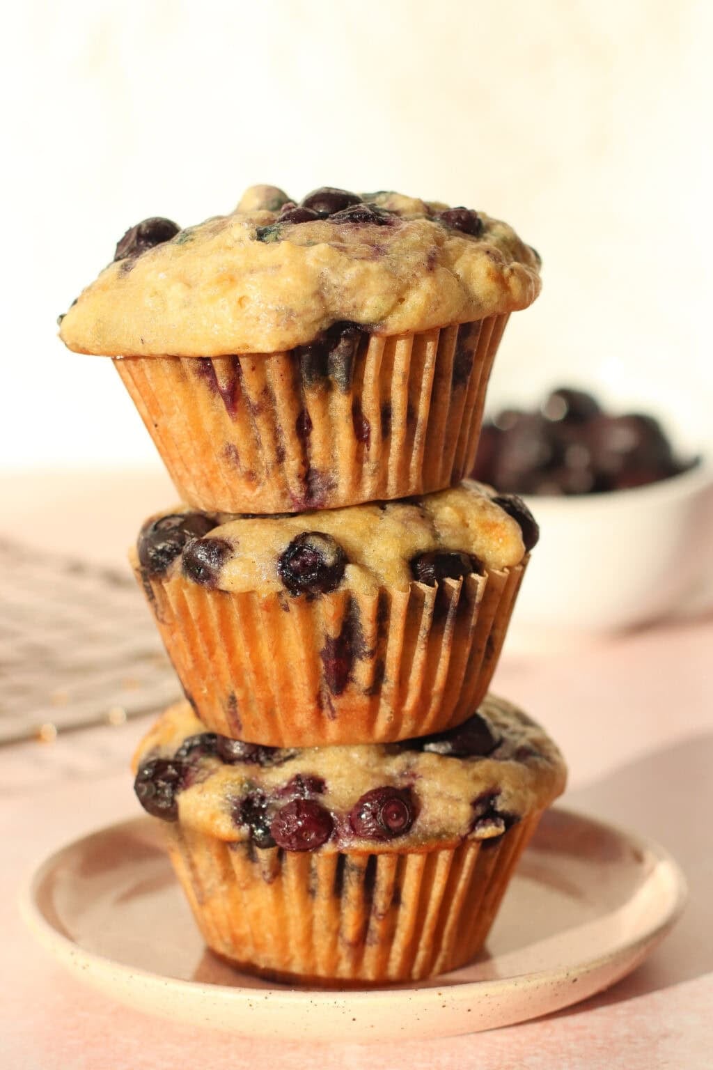 Three blueberry muffins are stacked on a round plate, with a bowl of blueberries and a cooling rack in the background, all set on a light pink surface.