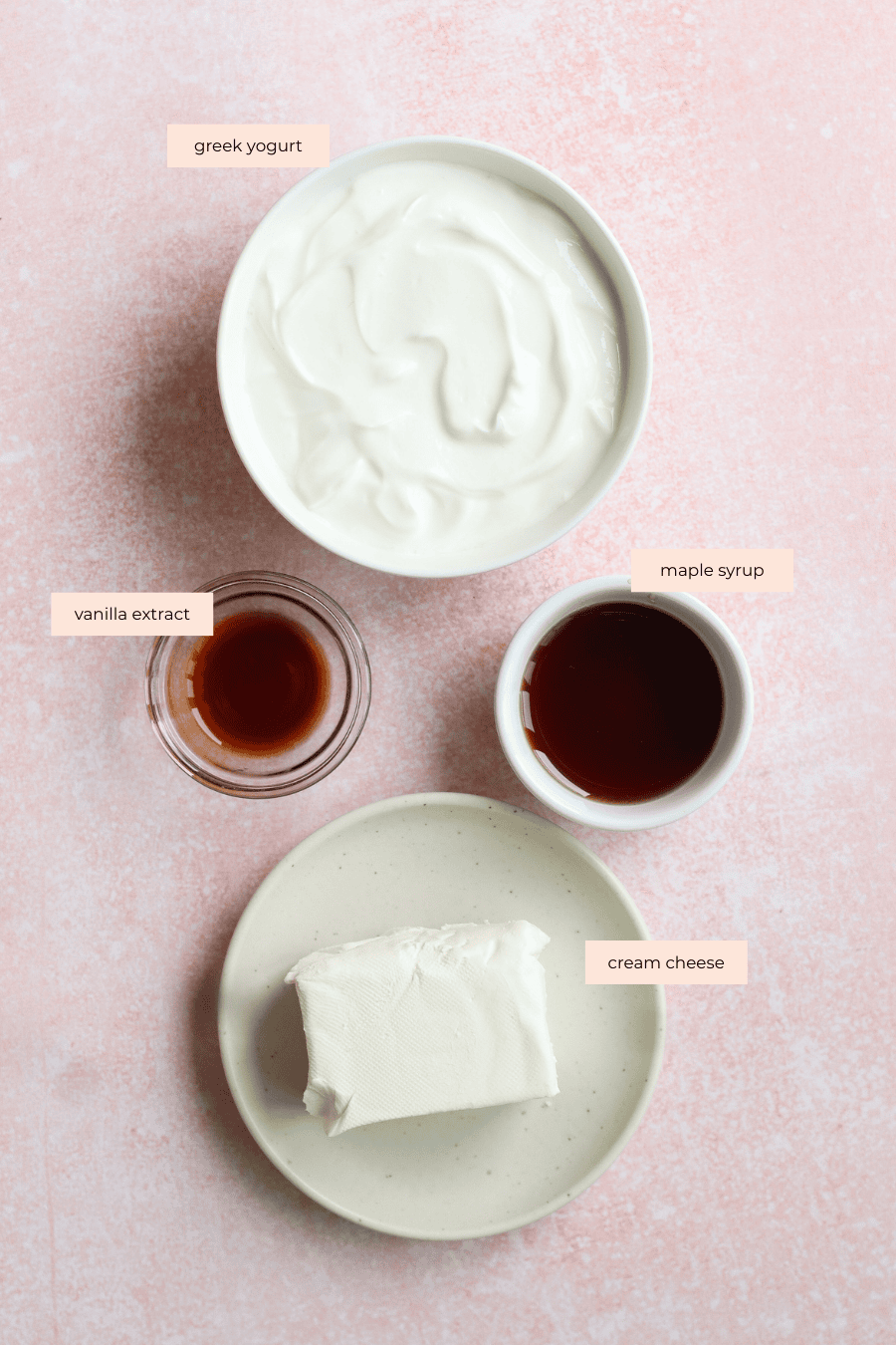 Four labeled ingredients on a pink surface: a bowl of Greek yogurt, a small glass of vanilla extract, a small bowl of maple syrup, and a plate with a block of cream cheese.