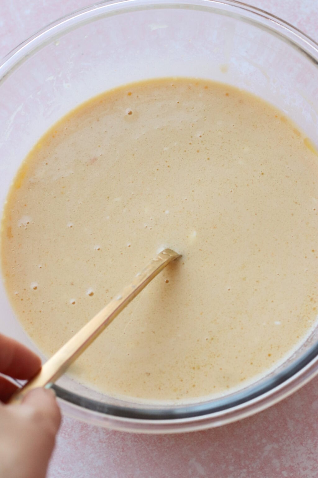 A hand is stirring a light tan batter in a clear glass bowl with a metal spoon, set on a pale pink surface.