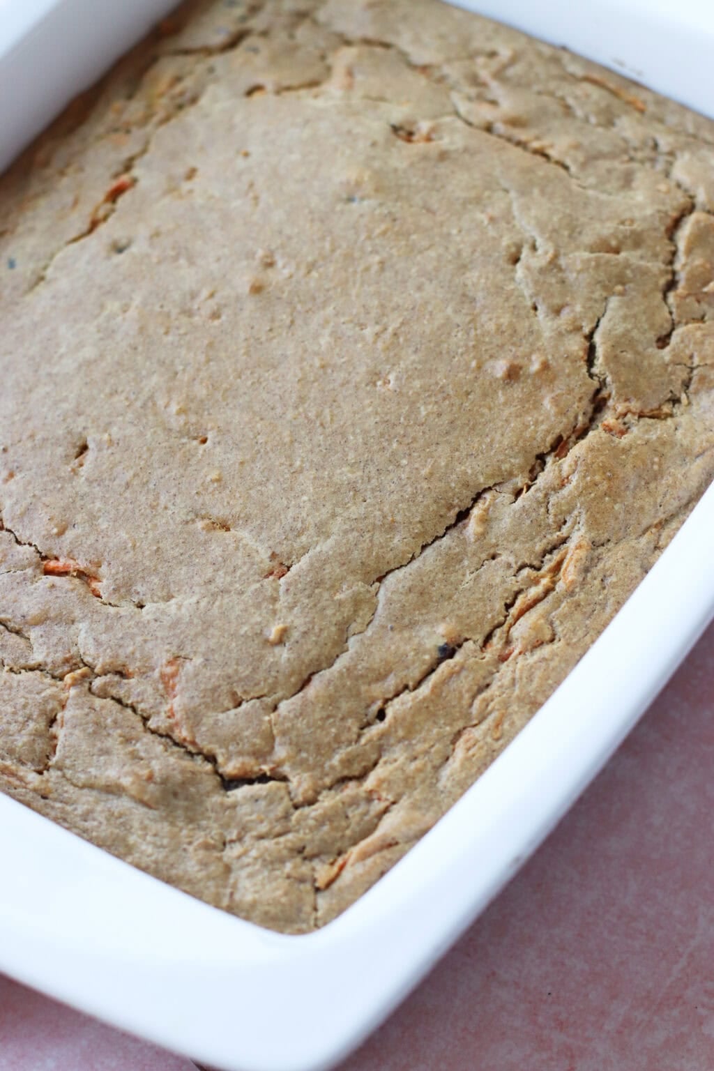A close-up of a freshly baked, golden-brown cake or bread in a white rectangular baking dish, showing a slightly cracked top surface.