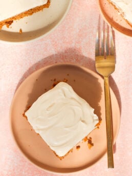 A square piece of cake with white frosting on a pink plate next to a gold fork, with another plate holding cake partially visible above. The background is light pink.