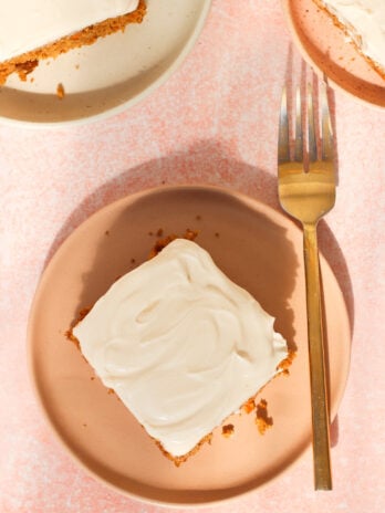 A square piece of cake with white frosting on a pink plate next to a gold fork, with another plate holding cake partially visible above. The background is light pink.