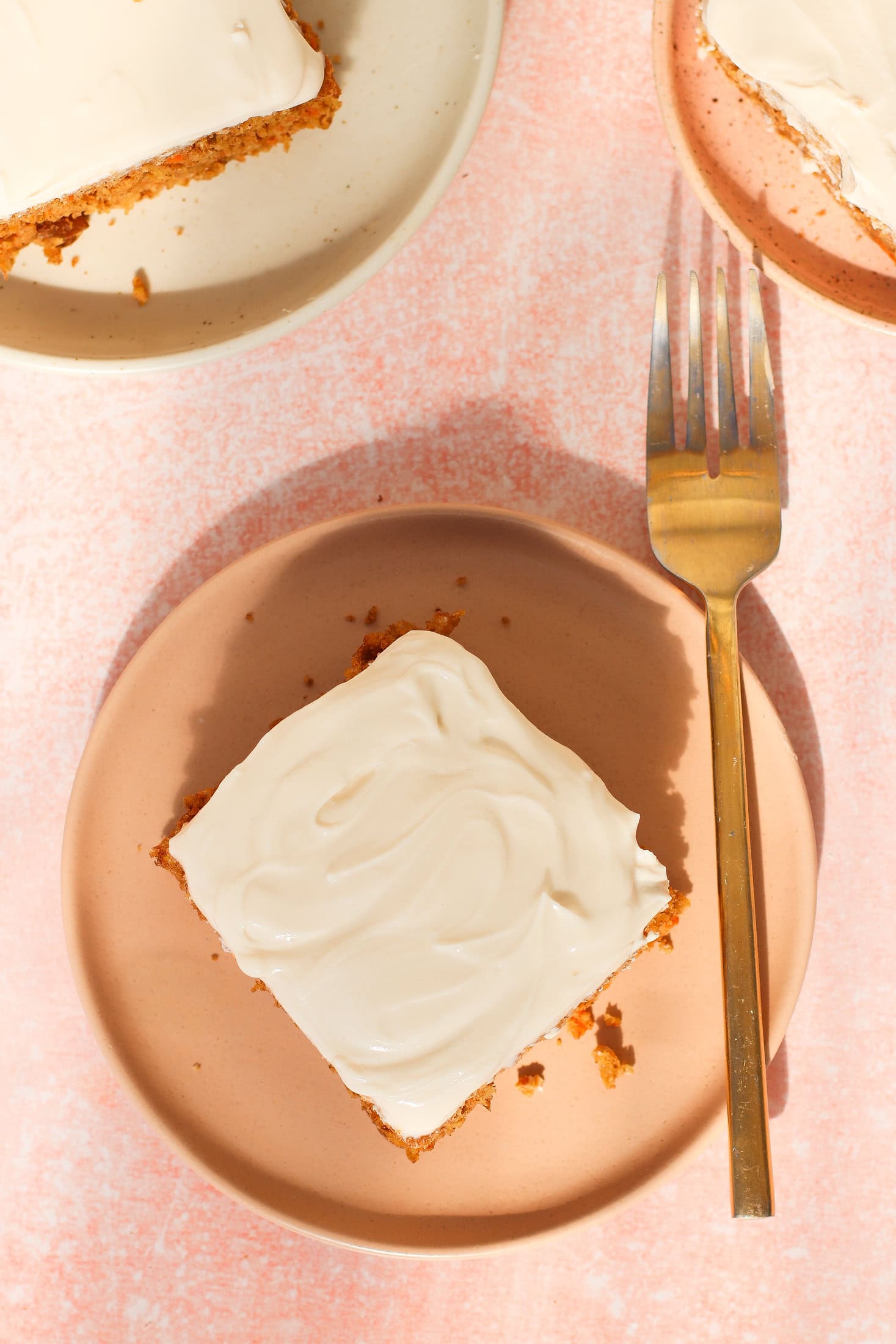 A square piece of cake with white frosting on a pink plate next to a gold fork, with another plate holding cake partially visible above. The background is light pink.