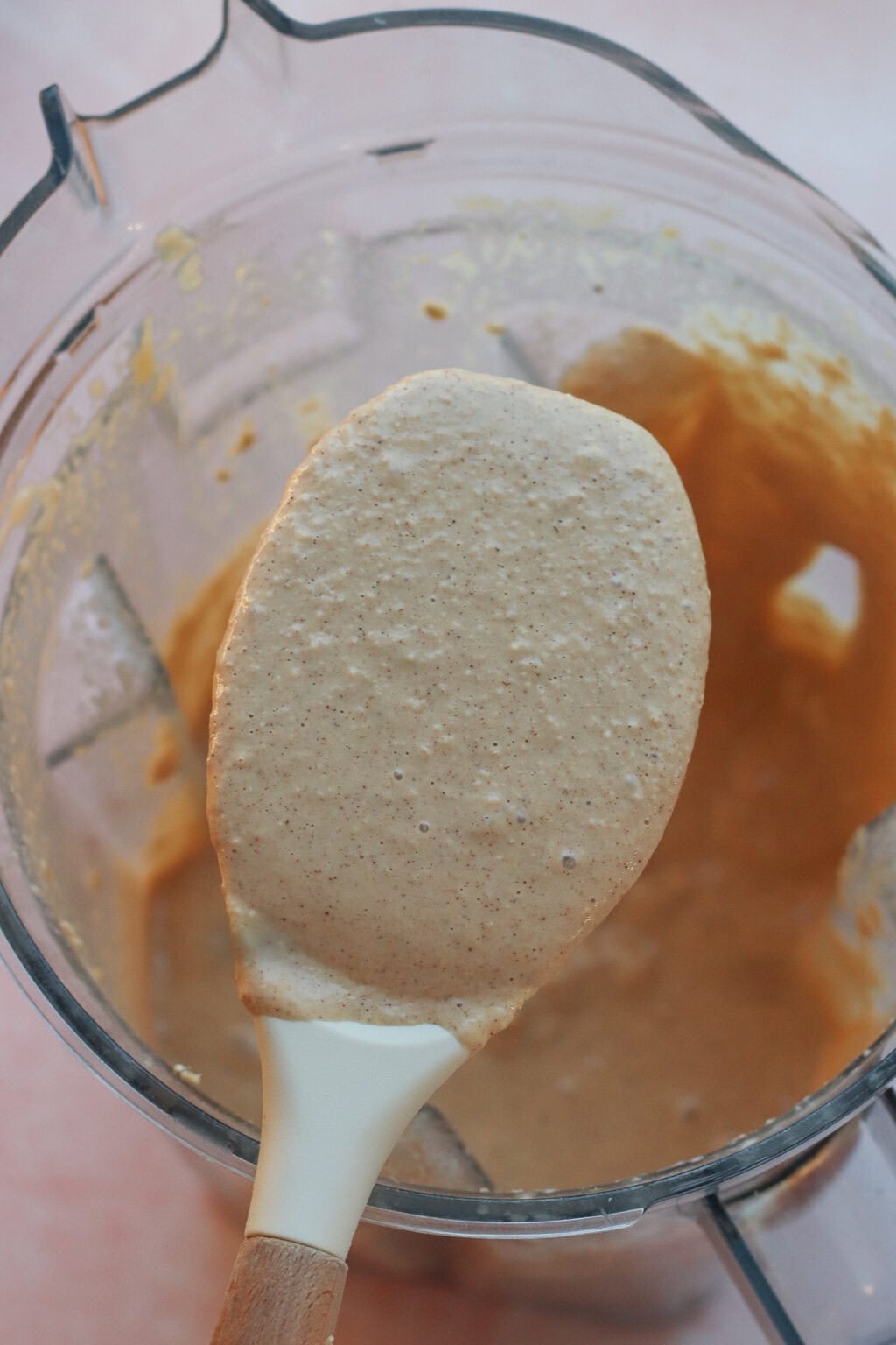 A close-up of a spatula covered in creamy, light brown batter held above a blender jar containing more of the same mixture. The background is softly blurred.