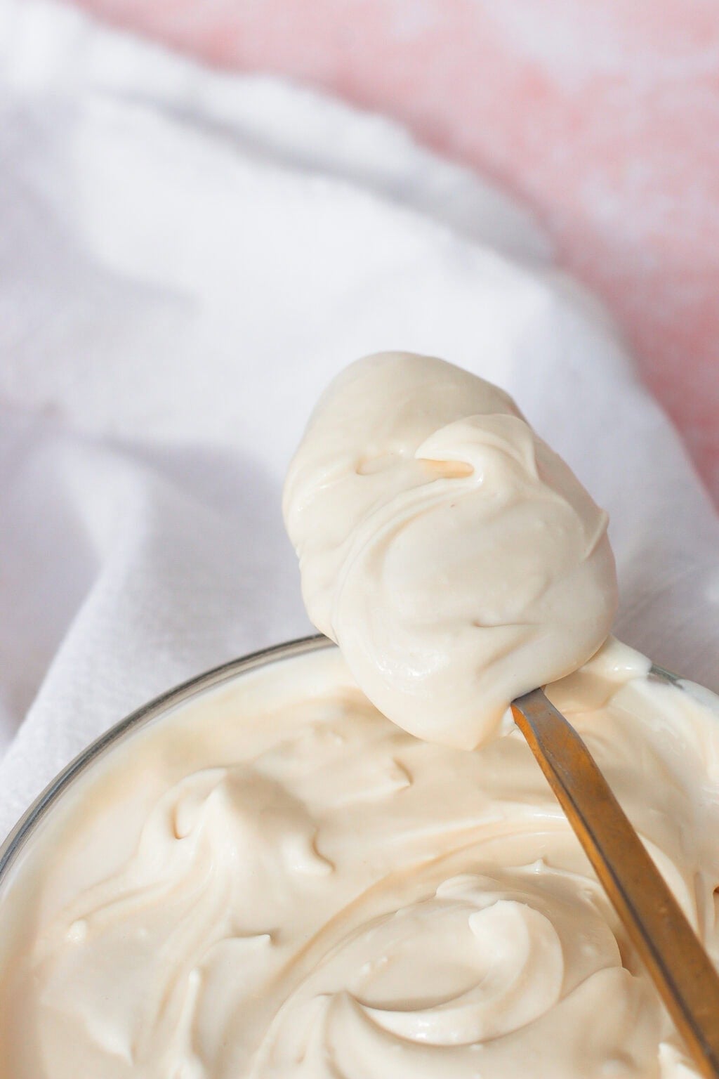 Close-up of a spoonful of creamy, white sauce or dip held above a bowl filled with the same mixture, with a soft white cloth in the background.