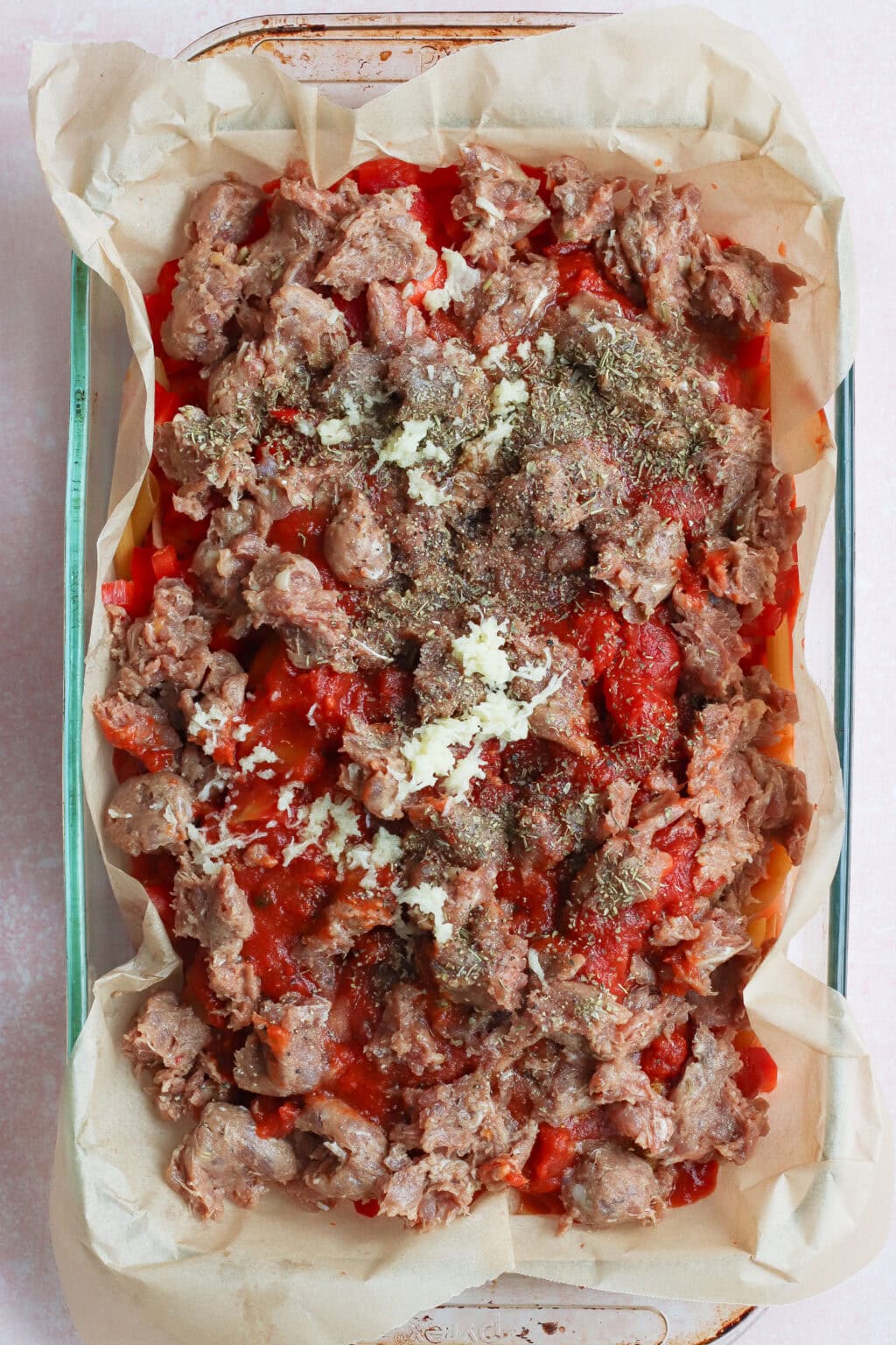 A glass baking dish lined with parchment paper, filled with layers of tomato sauce, crumbled sausage, garlic, and herbs, ready to be baked.
