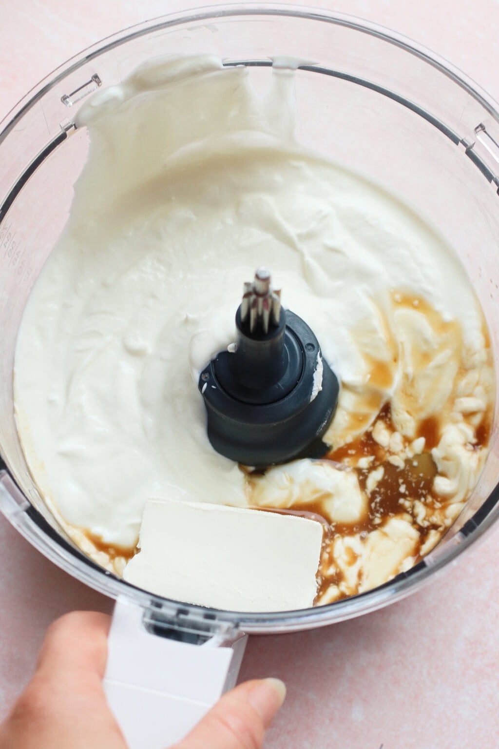 A food processor containing cream cheese, yogurt, vanilla extract, and a hand holding a spatula against the side, ready to blend the ingredients.