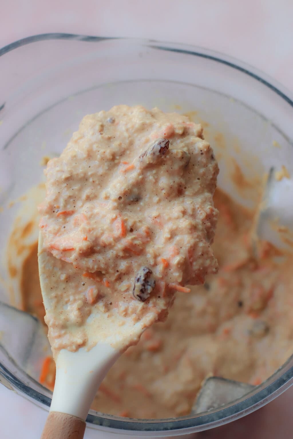 A close-up of a spatula lifting thick, textured batter with visible shredded carrots and raisins from a clear mixing bowl.