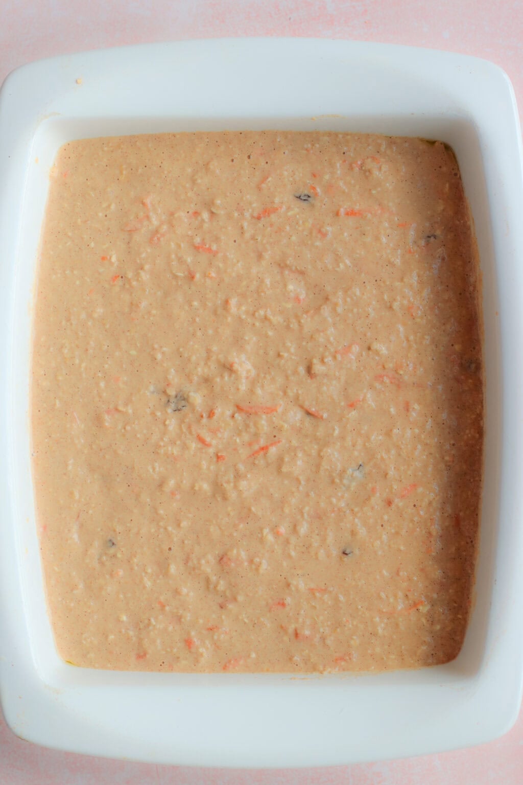 A square white baking dish filled with an uncooked, light brown batter that has visible specks and shreds, likely carrots and other ingredients, spread evenly across the surface.