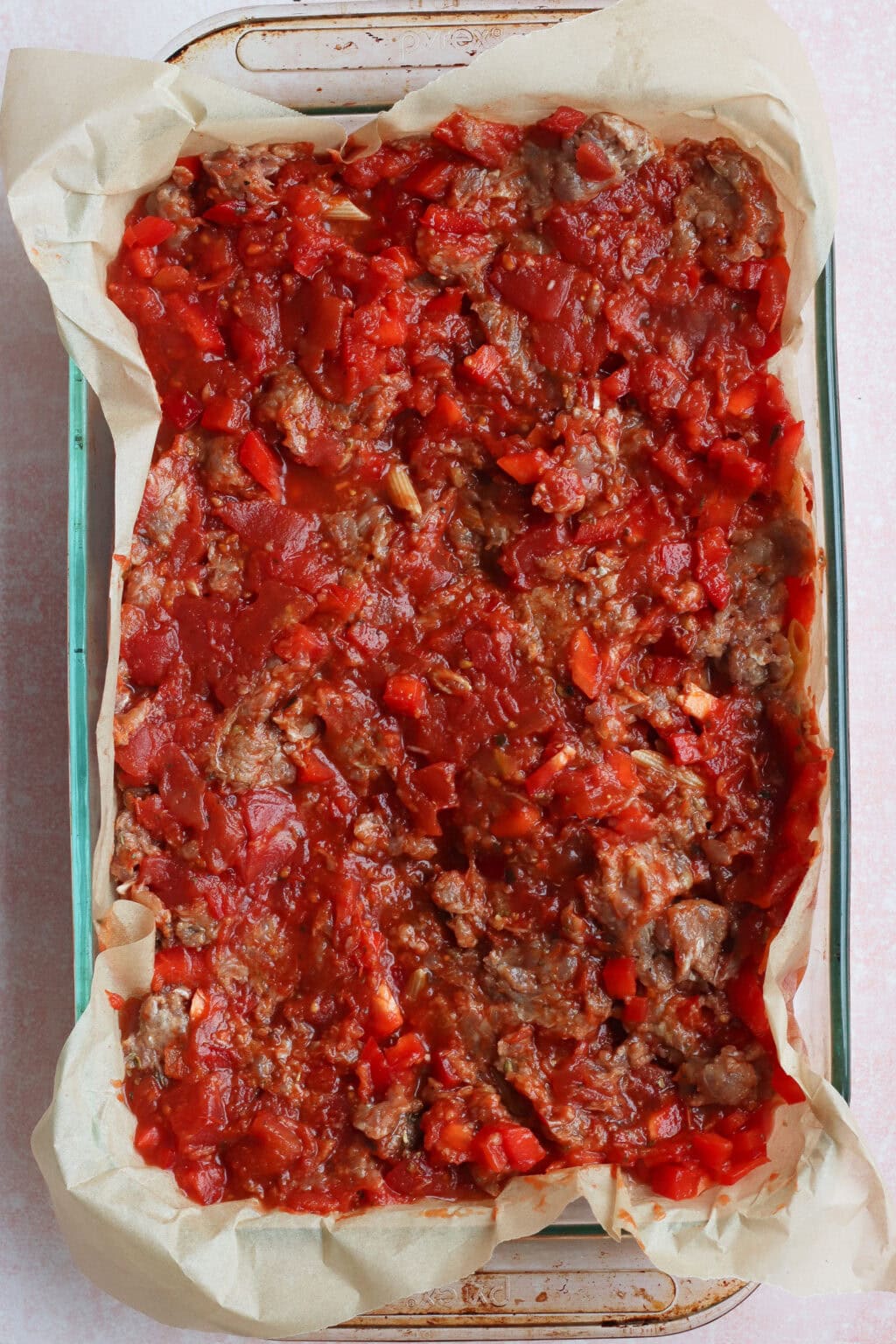 A glass baking dish lined with parchment paper is filled with a chunky tomato and ground meat mixture, spread evenly and ready to be baked.