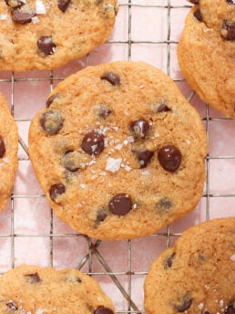 Chocolate chip cookies topped with flaky sea salt are cooling on a wire rack. The cookies are golden brown with visible chocolate chips and a light dusting of salt crystals. The background is a soft pink color.