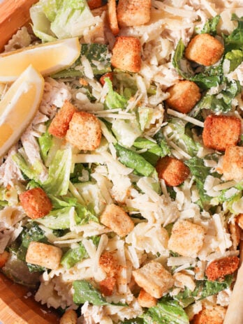 A close-up of Caesar salad with romaine lettuce, shredded cheese, seasoned croutons, lemon wedges, and grilled chicken in a wooden bowl.