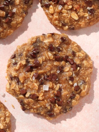 A close-up of a chewy chocolate chip peanut butter banana oatmeal cookie topped with sea salt flakes, resting on a light pink surface. The cookie is packed with oats, small chocolate chips, and hints of sweet banana flavor.