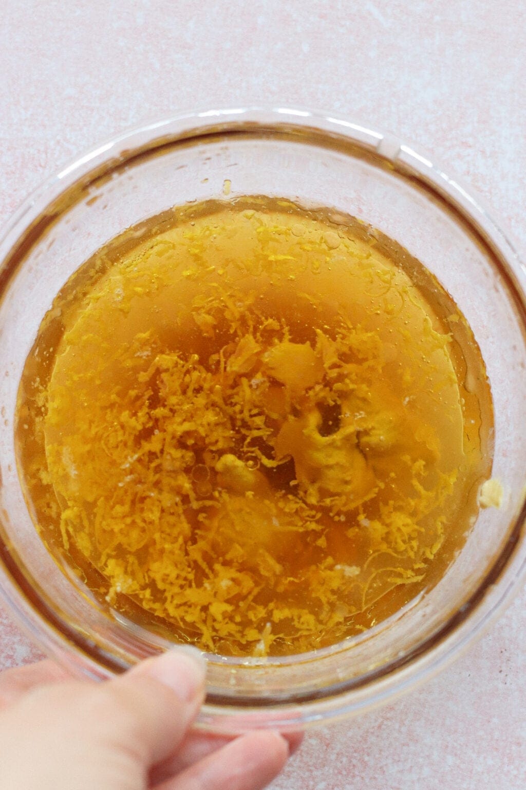 A hand holds a glass bowl filled with a golden-brown liquid and visible lemon zest on top, set against a light pink textured background.