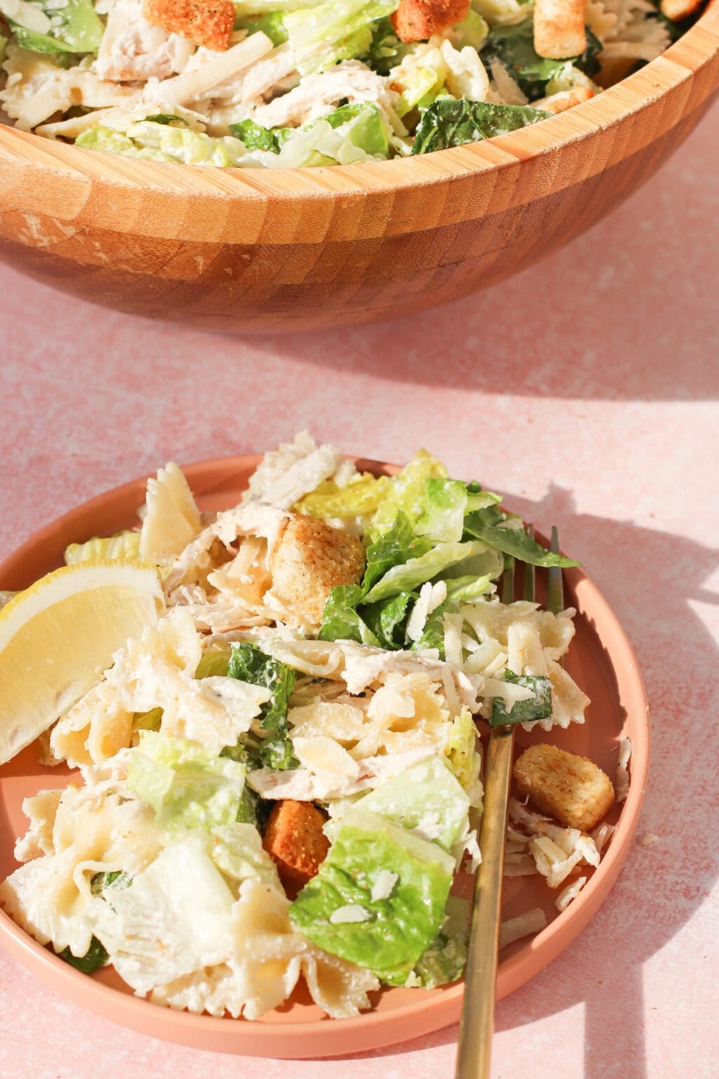 A plate of bowtie pasta salad with romaine lettuce, croutons, shredded chicken, and a lemon wedge sits next to a wooden bowl filled with more salad. A gold fork rests on the plate.