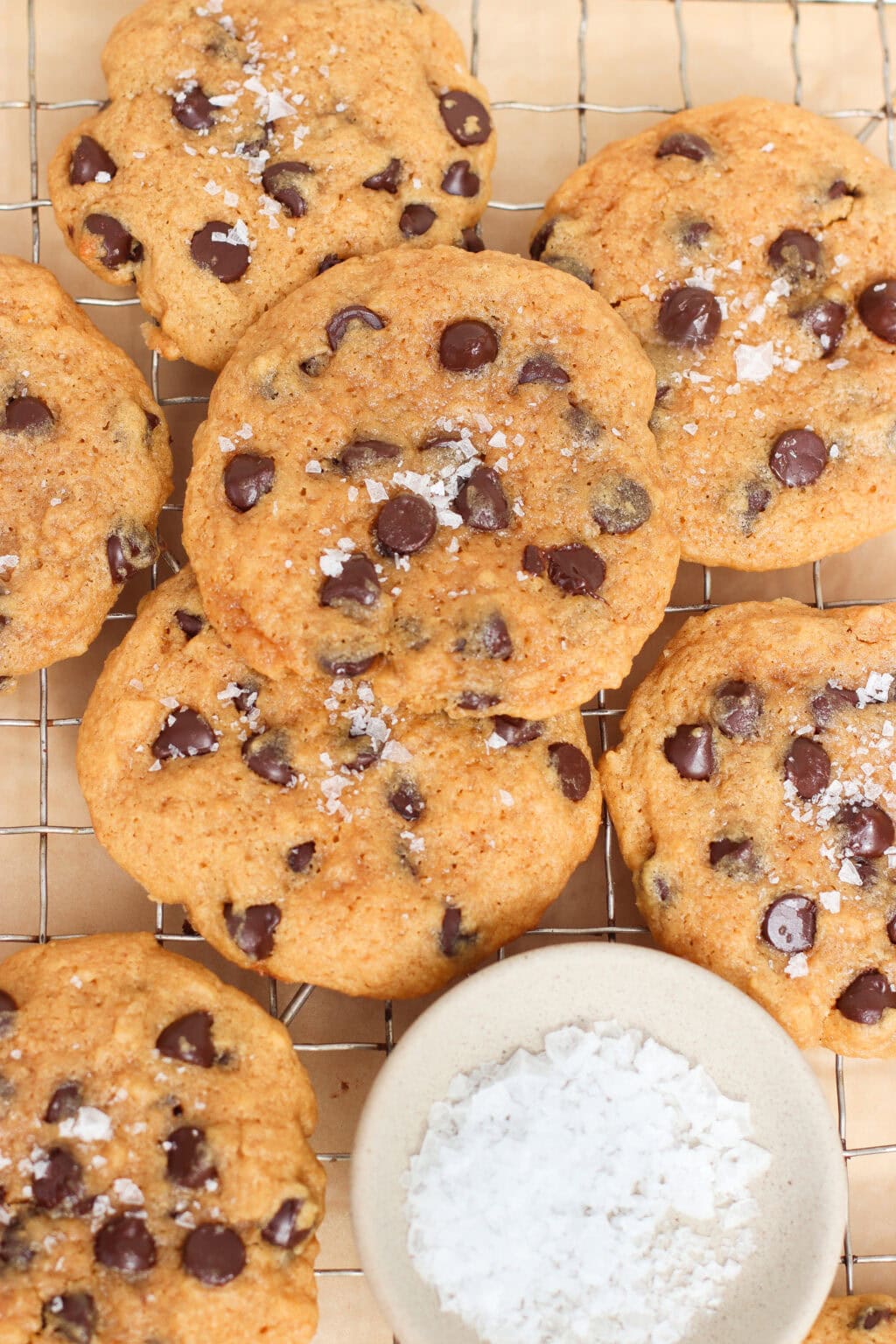 Chocolate chip cookies sprinkled with flaky sea salt are cooling on a wire rack next to a small bowl filled with coarse sea salt. The cookies look golden brown and freshly baked.