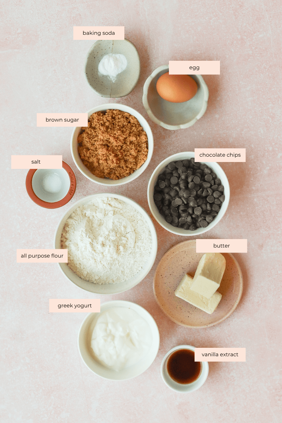 Top view of labeled baking ingredients for Greek yogurt chocolate chip cookies on a pink surface: baking soda, egg, brown sugar, chocolate chips, salt, flour, butter, Greek yogurt, and vanilla extract in small bowls or plates.