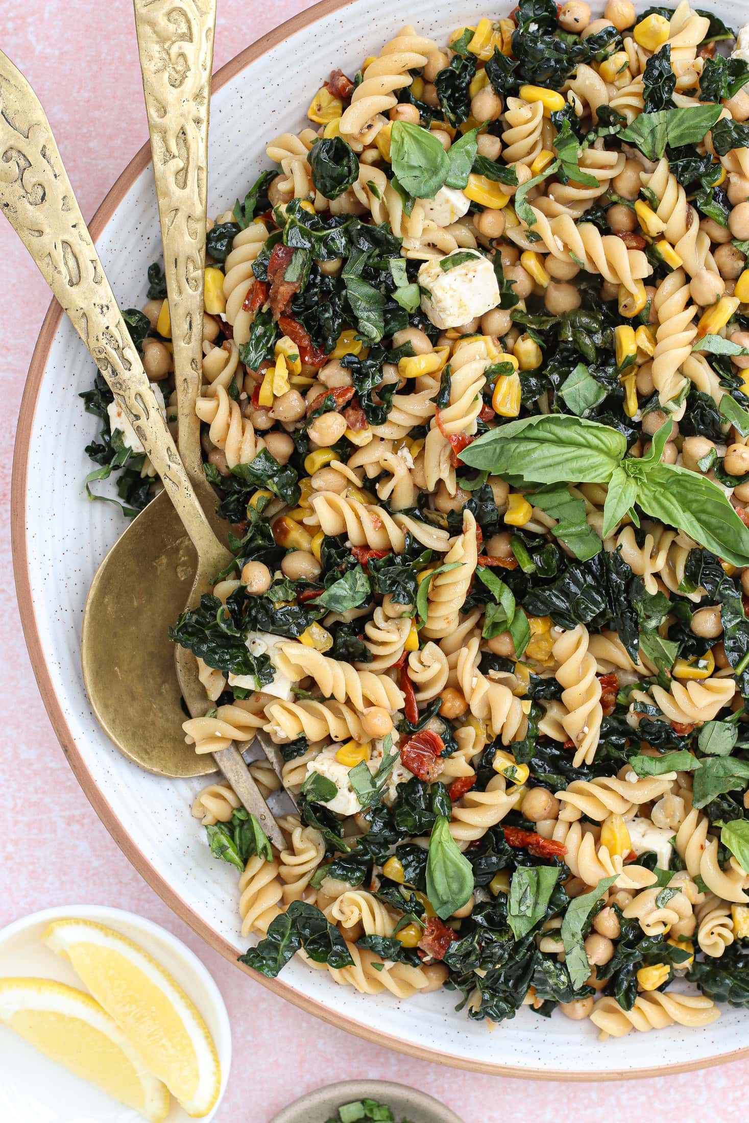 A large bowl of pasta salad with rotini, kale, chickpeas, sun-dried tomatoes, corn, fresh basil, and cheese, served with two ornate gold serving utensils. Lemon wedges are on the side.