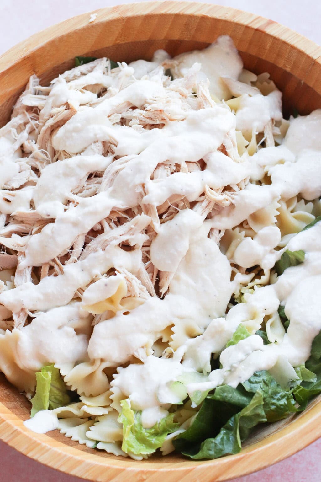 A wooden bowl filled with bow-tie pasta, shredded chicken, chopped romaine lettuce, and topped with a generous drizzle of creamy white dressing.