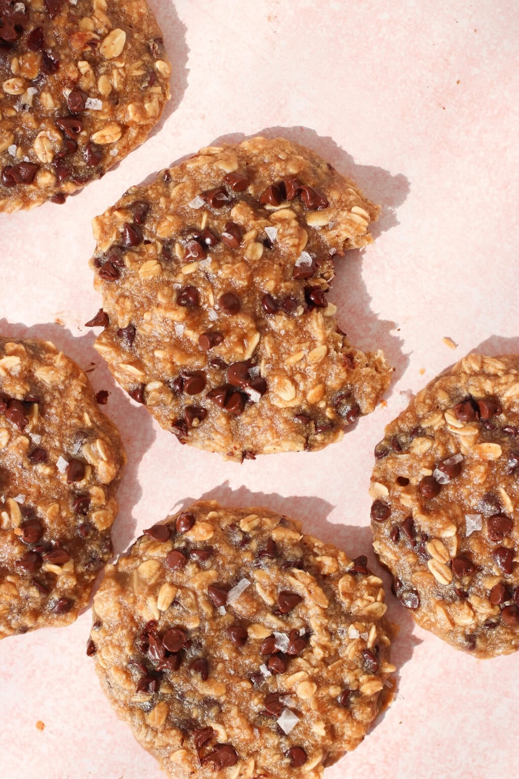 Five oatmeal chocolate chip cookies on a light pink surface, with one cookie missing a bite. The cookies are topped with chocolate chips and a sprinkle of flaky sea salt.