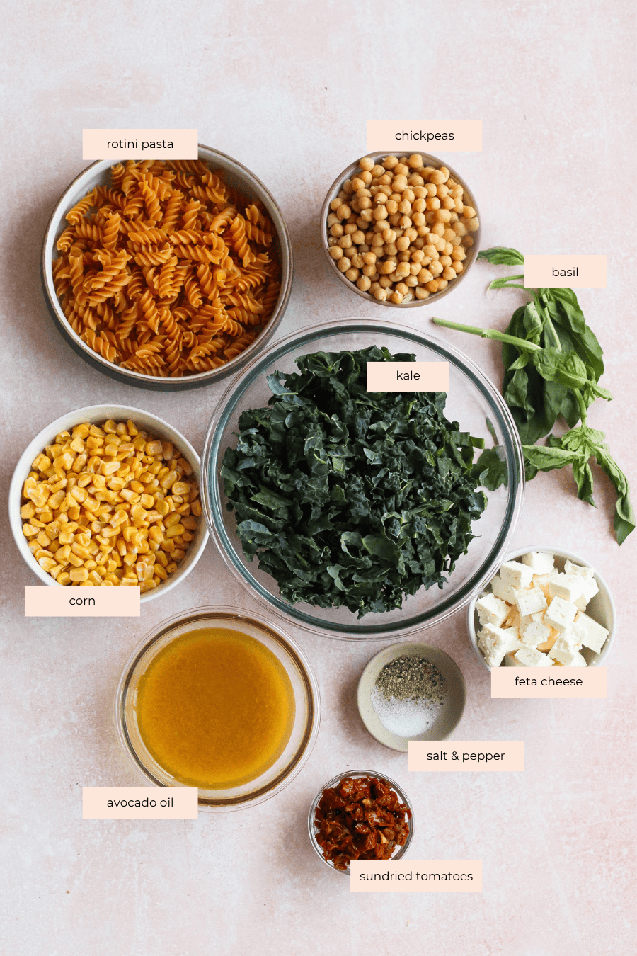 A flat lay of ingredients in bowls for a pasta salad: rotini pasta, chickpeas, fresh basil, chopped kale, corn, feta cheese, avocado oil, salt and pepper, and sundried tomatoes on a light pink background.