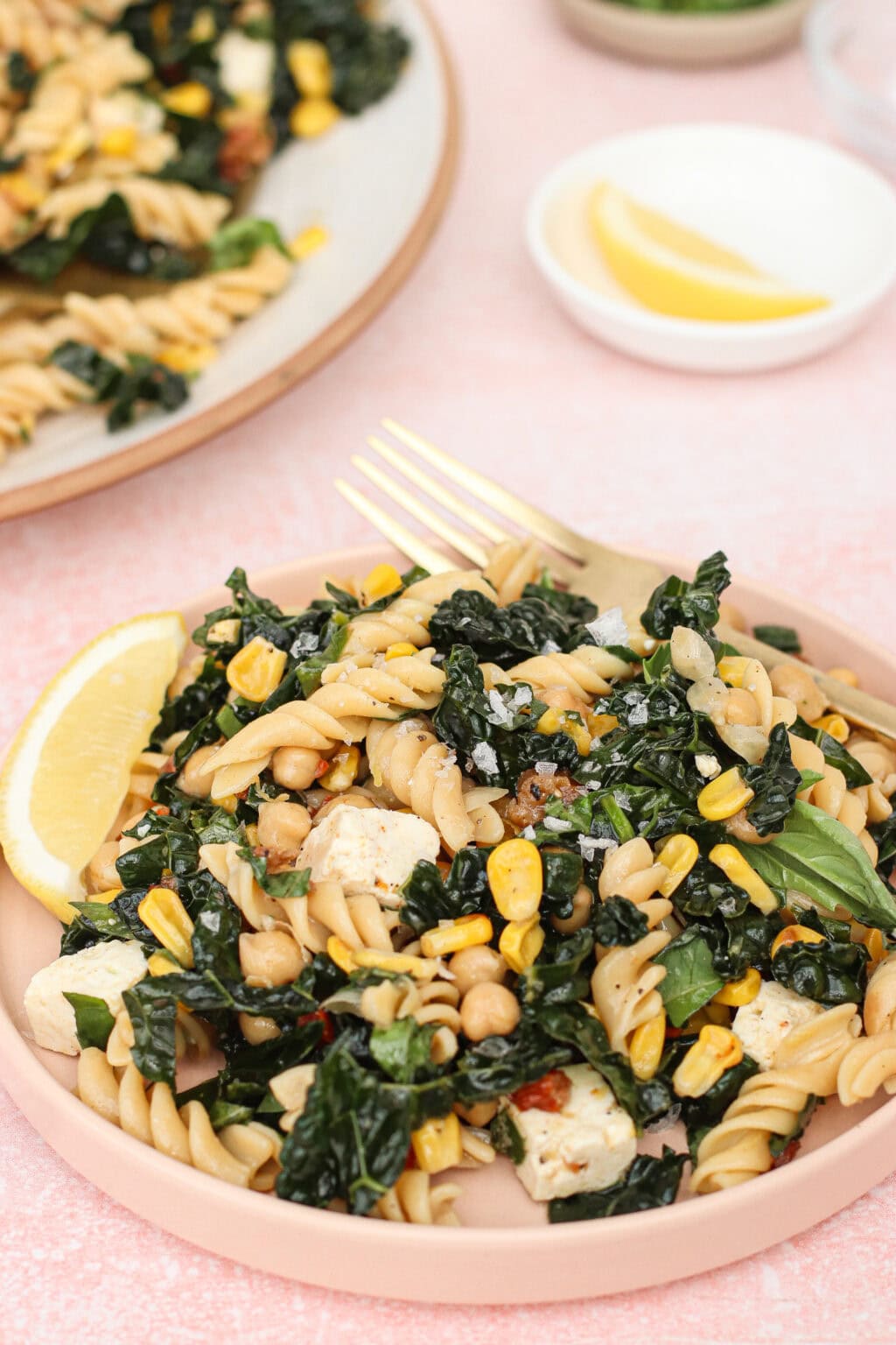 A plate of pasta salad with rotini, kale, corn, chickpeas, tofu, and herbs, garnished with a lemon wedge. A gold fork rests on the plate, and a small bowl with a lemon wedge is in the background.