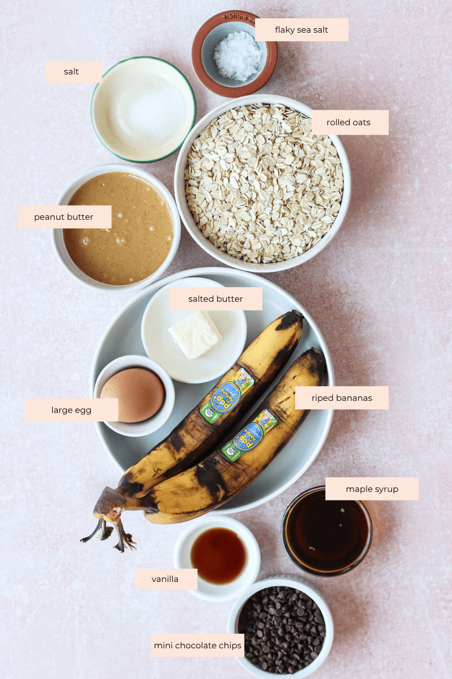 A flat lay of labeled baking ingredients: salt, flaky sea salt, rolled oats, peanut butter, salted butter, large egg, ripe bananas, maple syrup, vanilla, and mini chocolate chips on a light pink surface.