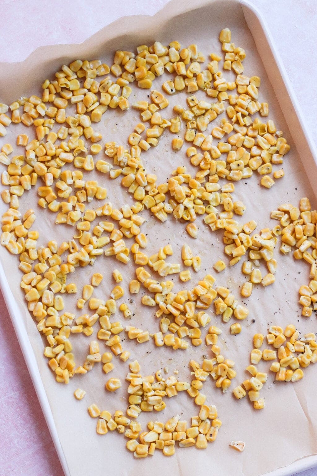 A baking tray lined with parchment paper, scattered with roasted corn kernels seasoned with pepper and spices, ready to be baked or already lightly roasted.