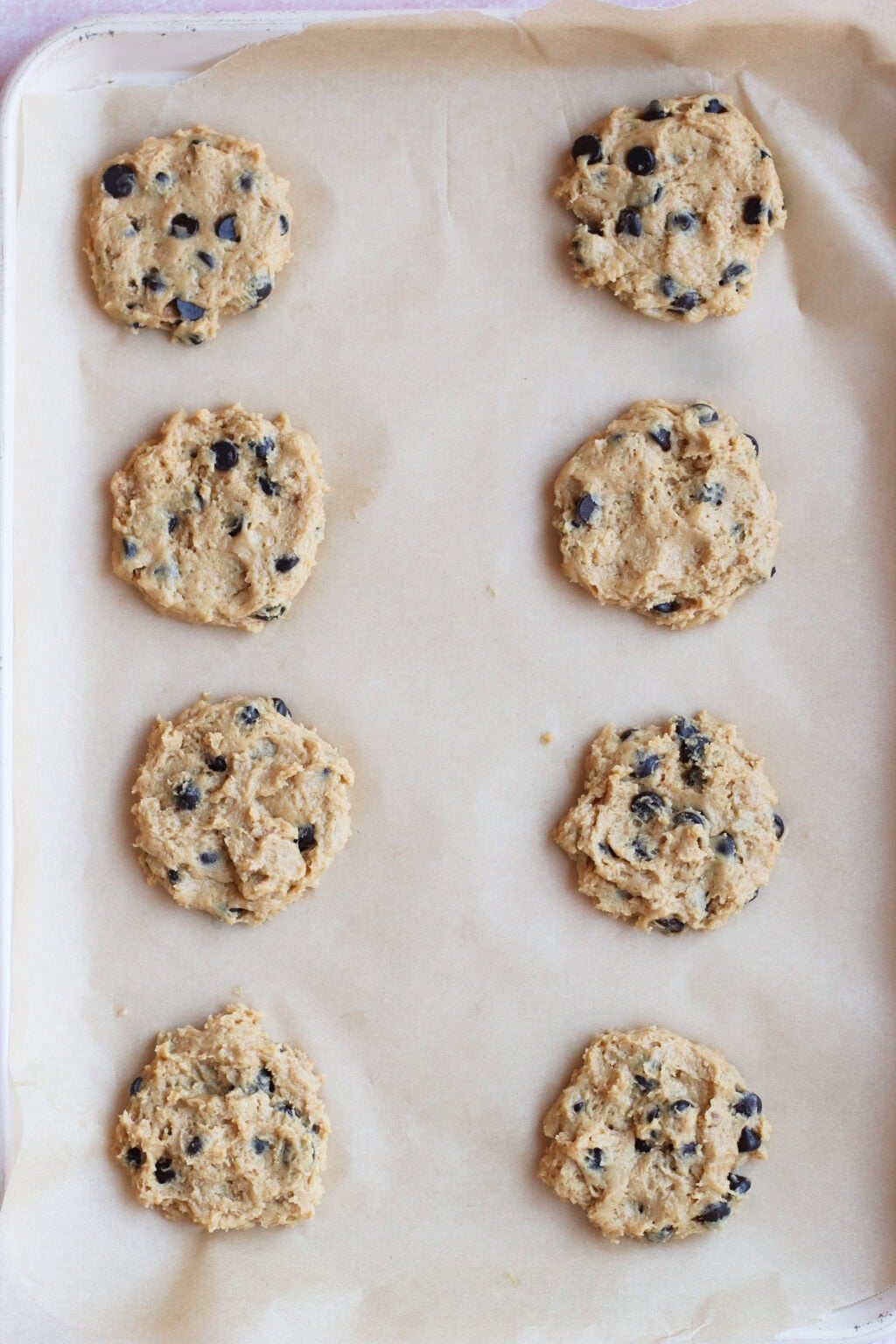 Eight raw chocolate chip cookie dough mounds are spaced out on a parchment-lined baking sheet, ready to be baked.