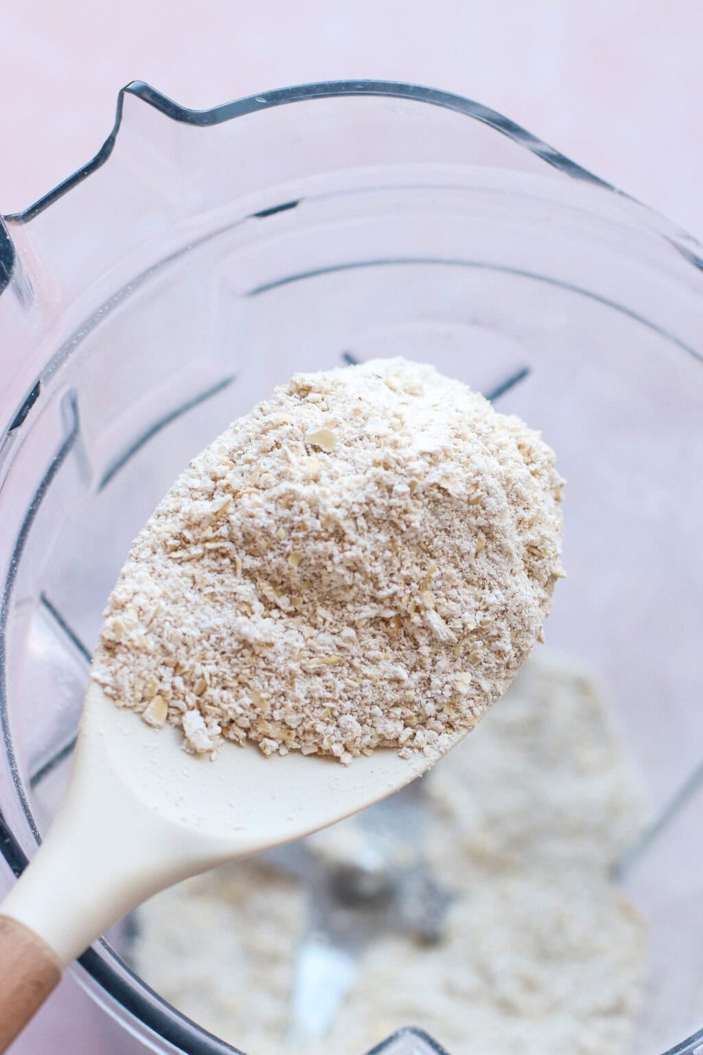 A close-up of a white spatula holding finely ground oat flour over a blender bowl filled with more oat flour. The background is soft pink, and the texture of the blended oats is visible.
