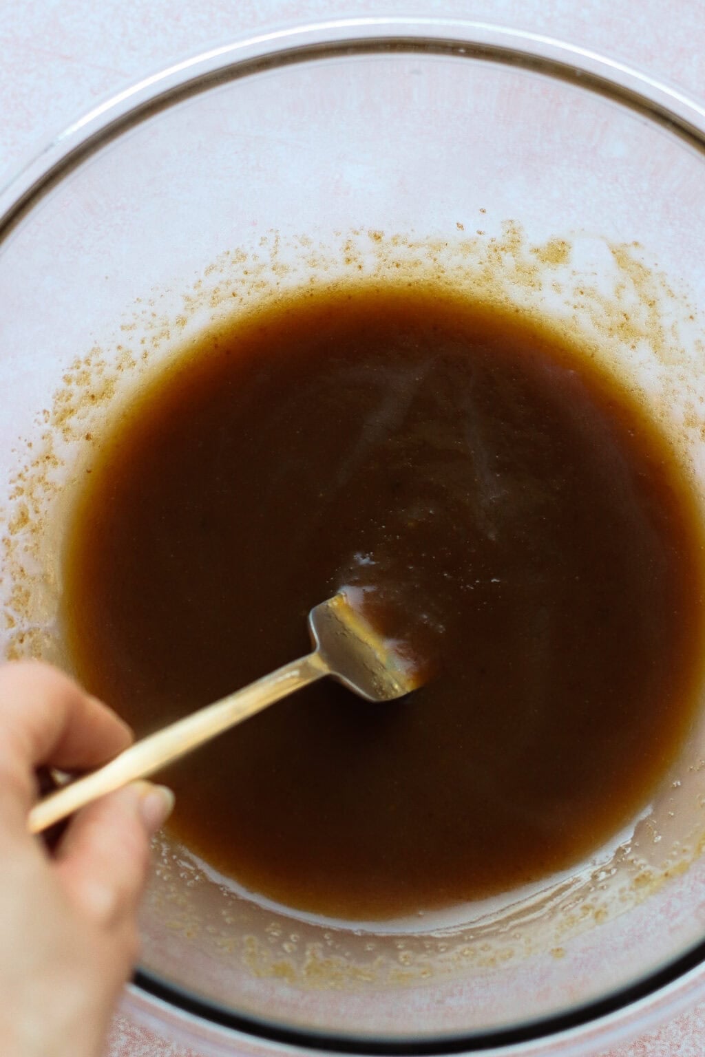 A hand holding a fork is stirring a thick, brown liquid in a large glass mixing bowl on a light-colored surface.