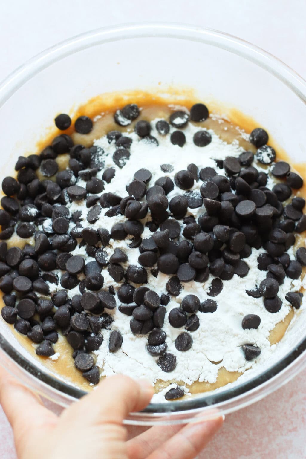 A hand holds a clear bowl filled with cookie dough, topped with chocolate chips and flour, ready to be mixed.