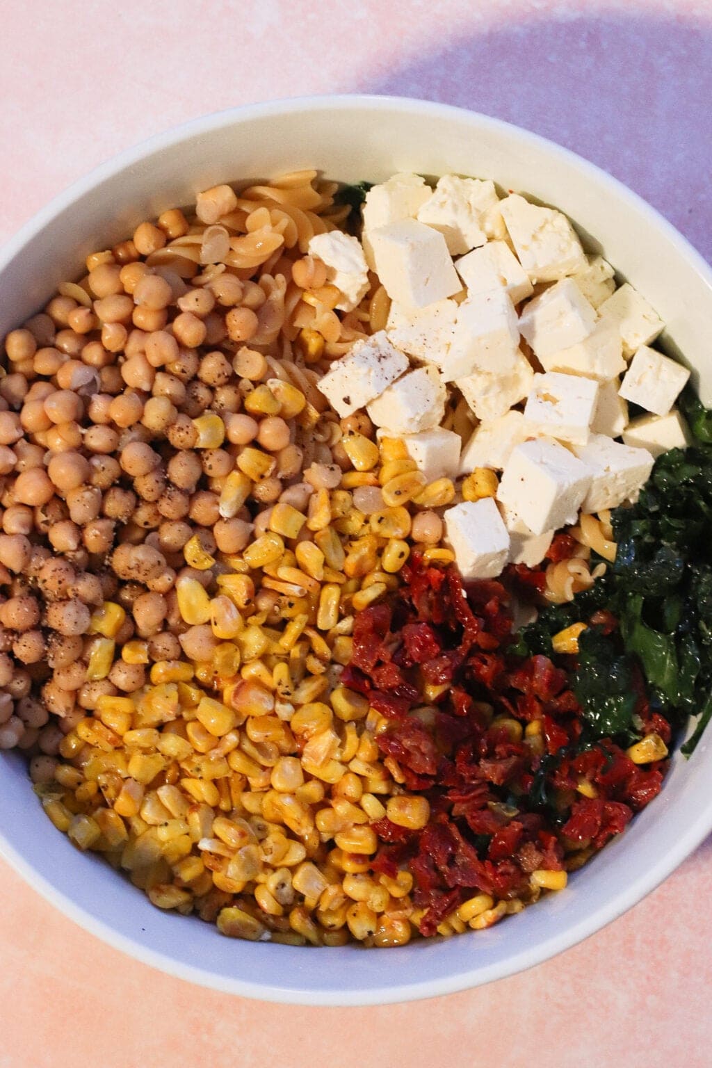 A bowl filled with chickpeas, corn, cubed tofu, chopped sun-dried tomatoes, wilted greens, and pasta, arranged in separate sections on a light pink surface.