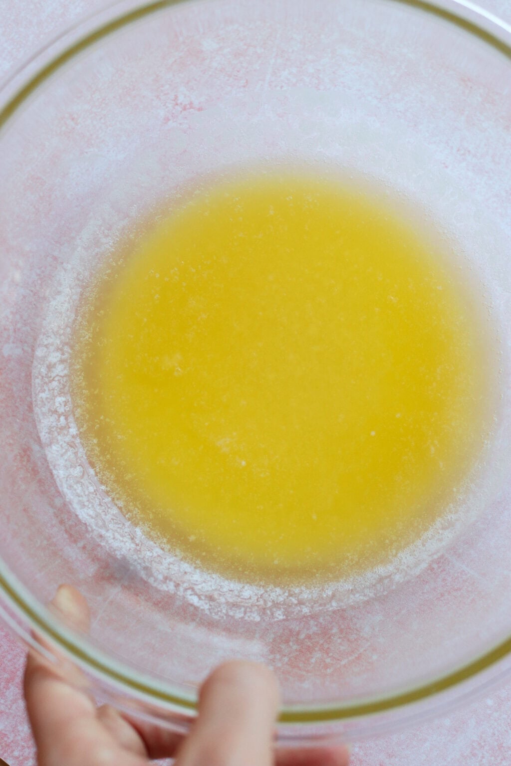 A hand holds a clear glass bowl filled with melted yellow butter, viewed from above on a light pink surface.