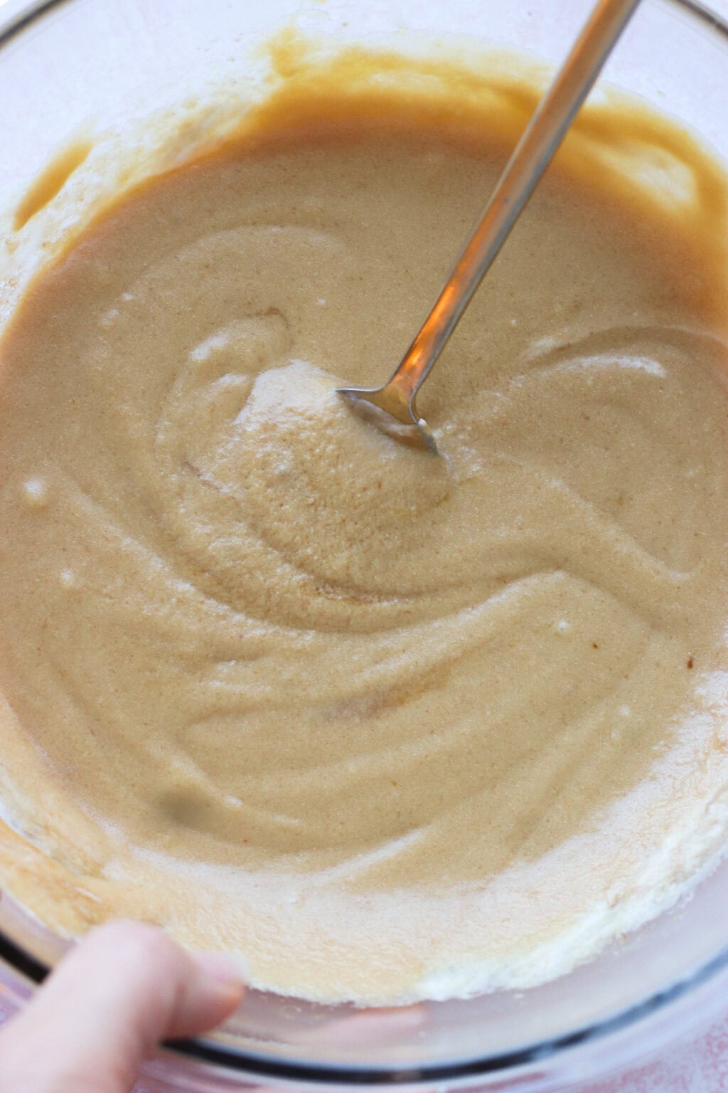 A close-up of a bowl filled with thick, creamy batter being mixed with a metal spoon. A hand is holding the edge of the bowl. The batter appears smooth and light brown in color.