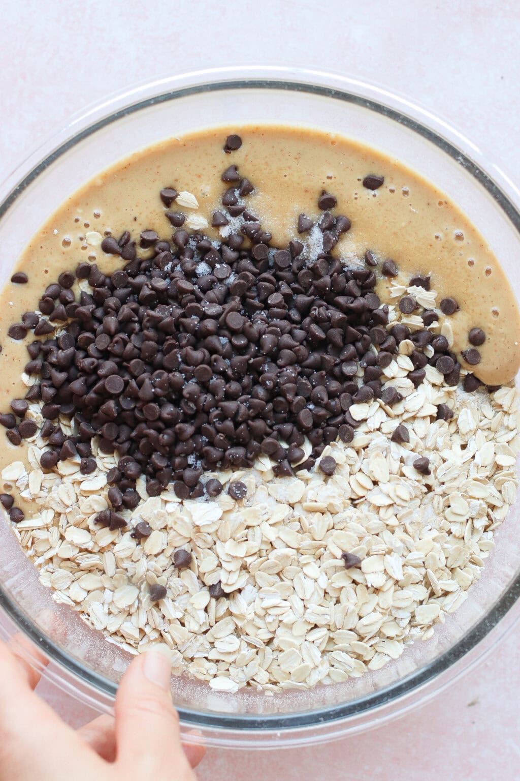 A glass bowl containing a mixture of oats, chocolate chips, and a light brown batter. A hand is holding the side of the bowl, preparing to mix the ingredients together.