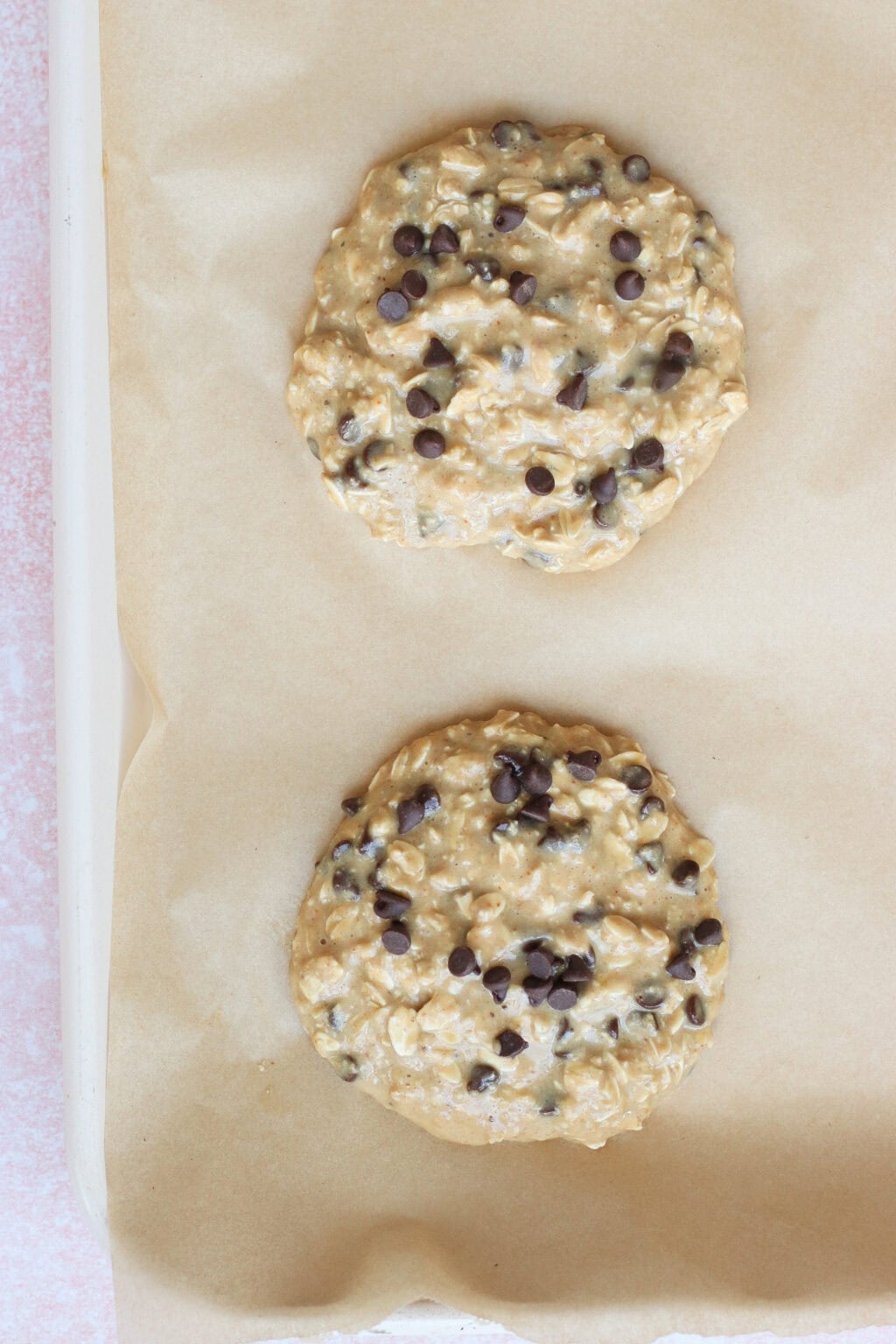 Two unbaked oatmeal chocolate chip cookies with visible chocolate chips sit on a parchment-lined baking sheet, ready to go into the oven.