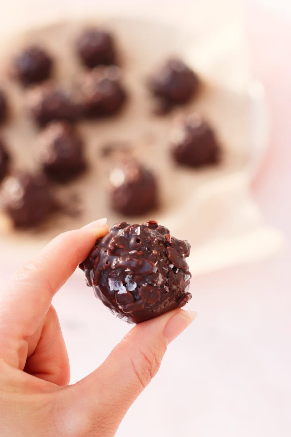 A hand holding a round, chocolate-covered treat with a textured surface; more similar treats are blurred in the background on parchment paper.
