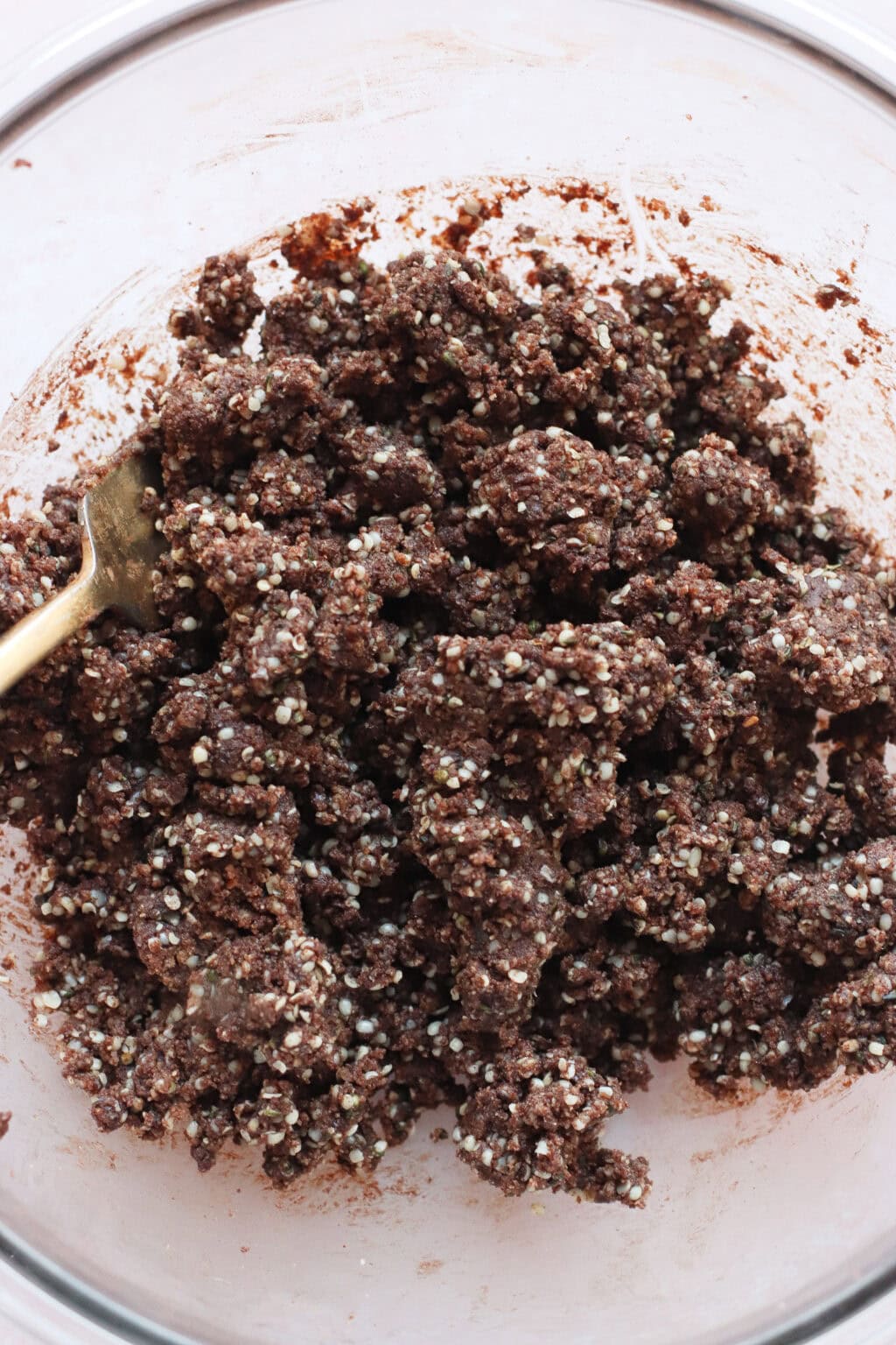 A close-up of a glass mixing bowl containing a thick, chunky mixture of chocolate and puffed grains, with a spoon partially visible on the left side.