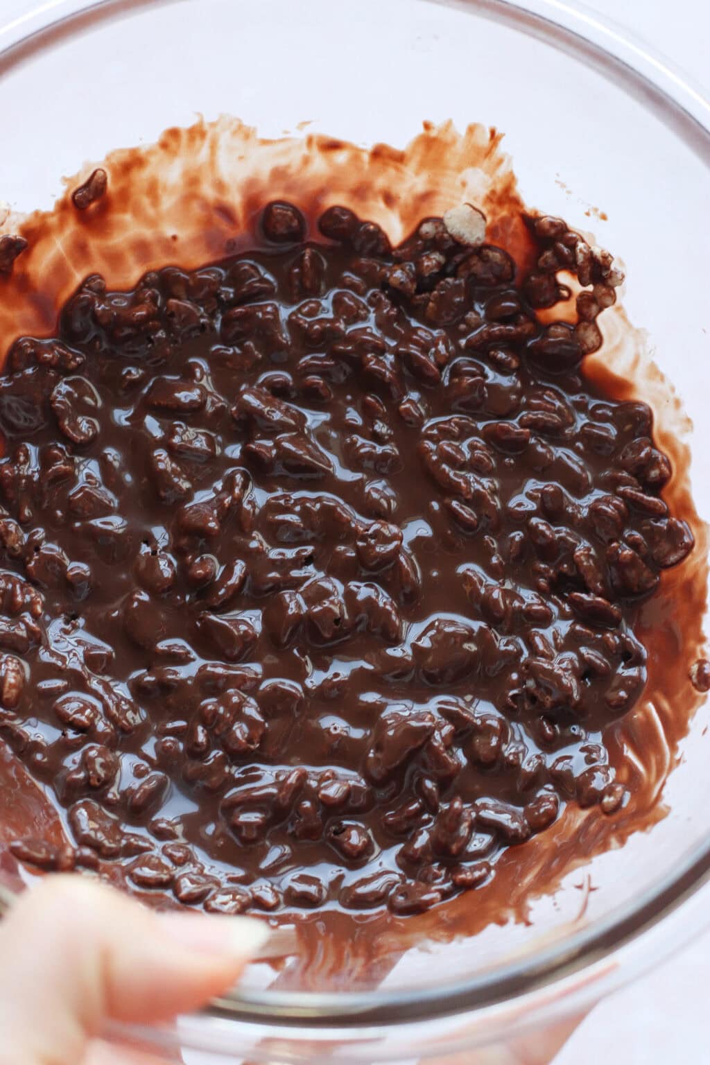 A glass bowl filled with a mixture of melted chocolate and crispy rice cereal, partially stirred together, with some chocolate smeared along the sides of the bowl.