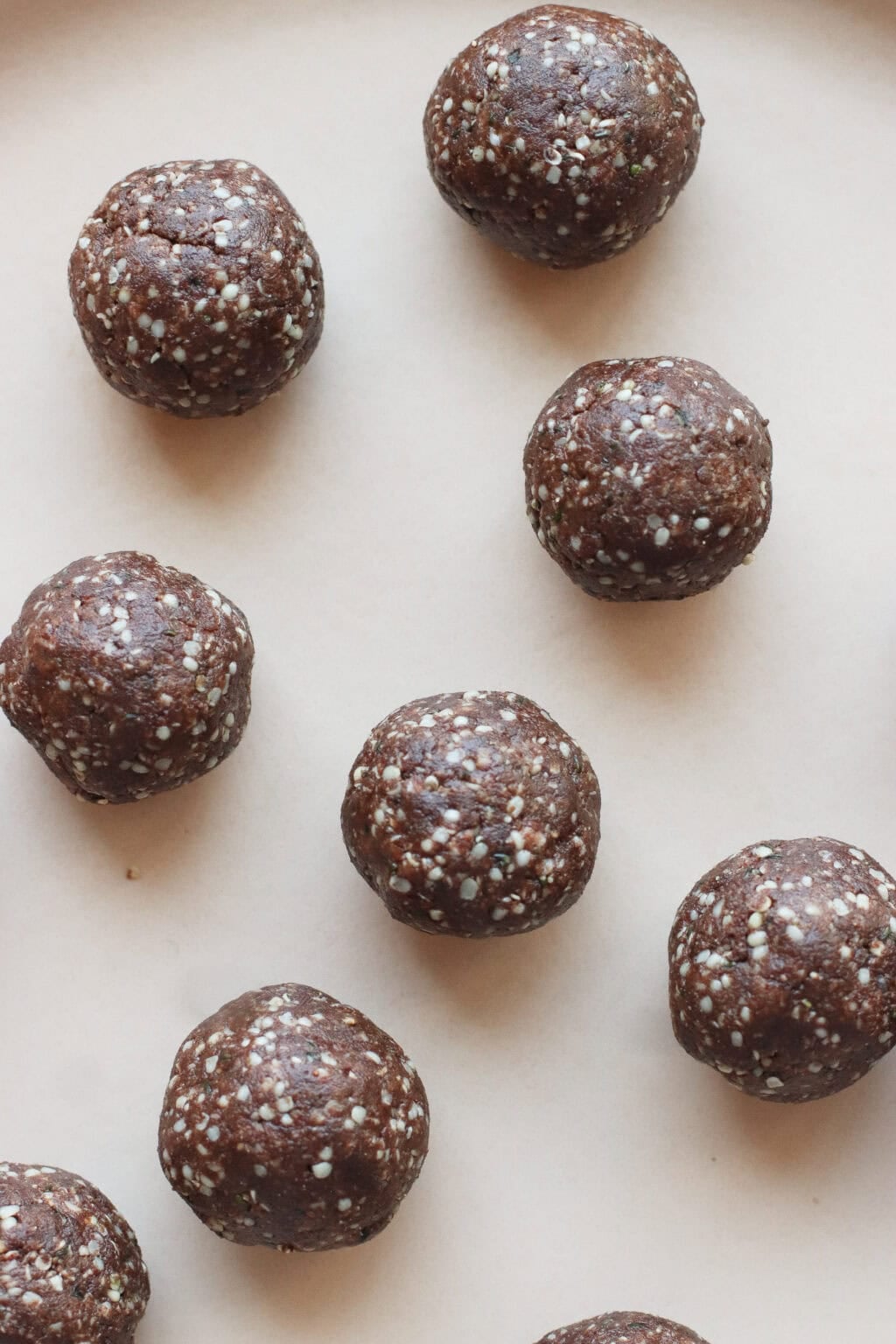 Nine chocolate energy balls with visible white seeds are arranged on a light beige surface. The balls have a rough texture and are evenly spaced across the image.