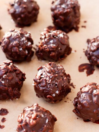 Chocolate-covered crunchy nut clusters arranged on parchment paper, with a glossy coating and a sprinkle of sea salt on top.