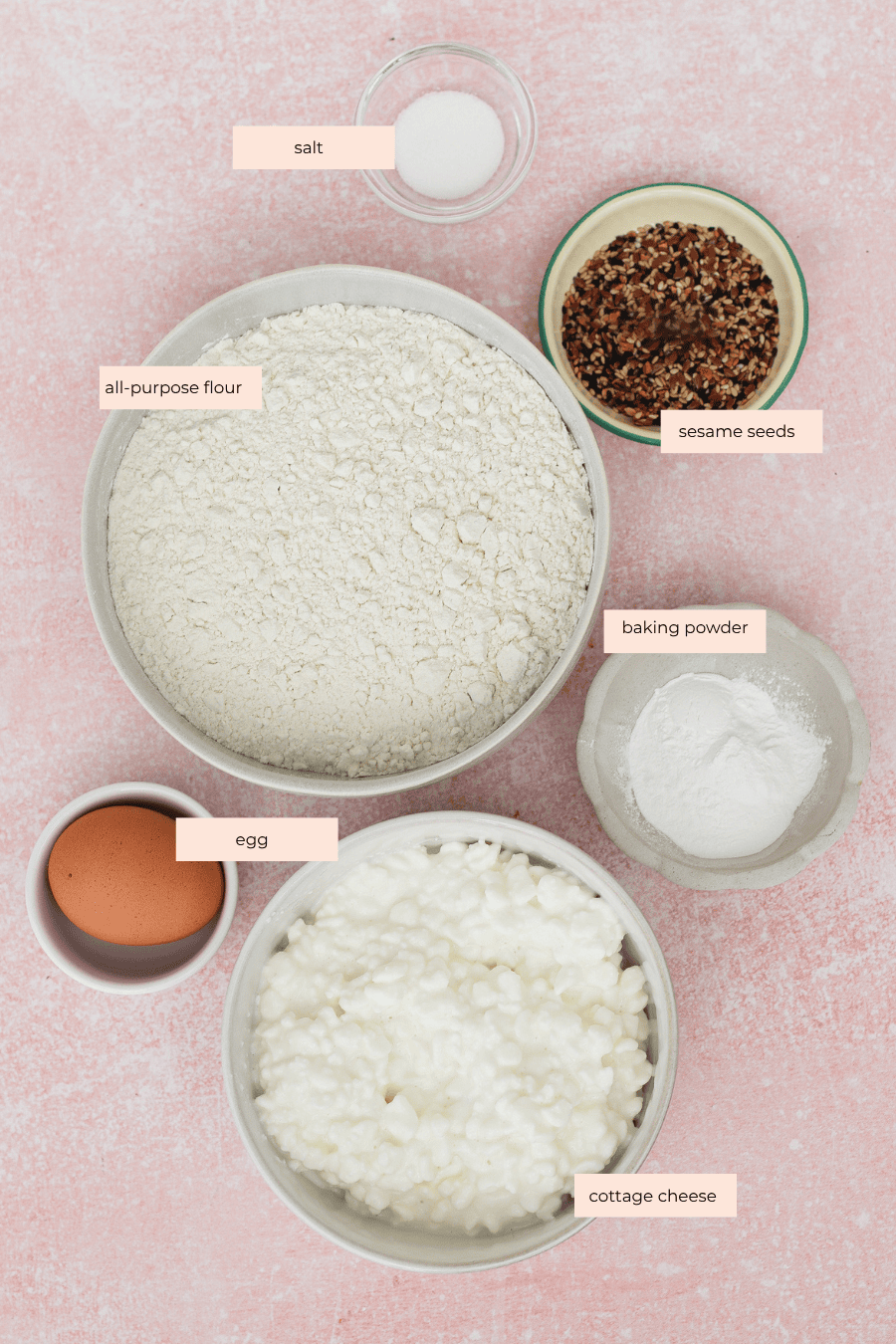 Six labeled bowls on a pink surface containing salt, all-purpose flour, sesame seeds, baking powder, an egg, and cottage cheese, arranged neatly for a recipe preparation.