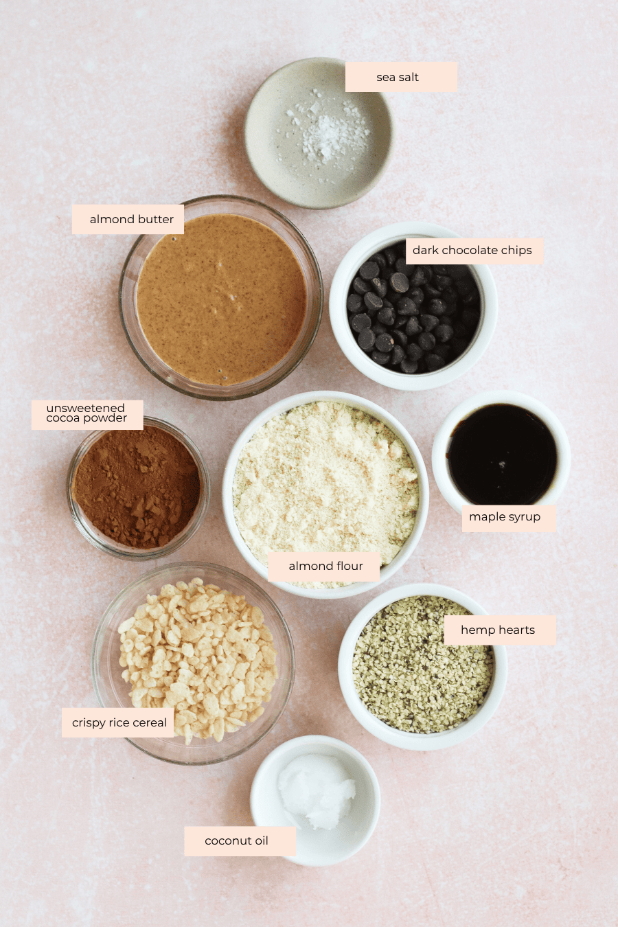 A flat lay of bowls containing sea salt, almond butter, dark chocolate chips, unsweetened cocoa powder, almond flour, maple syrup, crispy rice cereal, hemp hearts, and coconut oil on a light pink surface. Labels identify each ingredient.