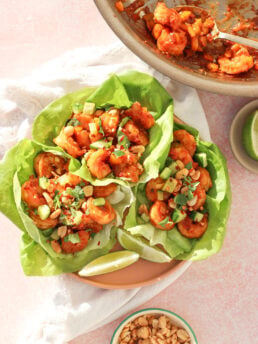Three lettuce wraps filled with saucy shrimp, diced avocado, chopped peanuts, and herbs are arranged on a plate. A bowl of extra peanuts and a pan with remaining shrimp are nearby. Lime wedges are on the plate.