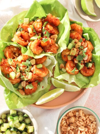Three lettuce wraps filled with spicy shrimp, chopped avocado, peanuts, and herbs are displayed on a plate, with lime wedges on the side. Bowls of diced cucumber and peanuts are nearby. Bright and fresh presentation.