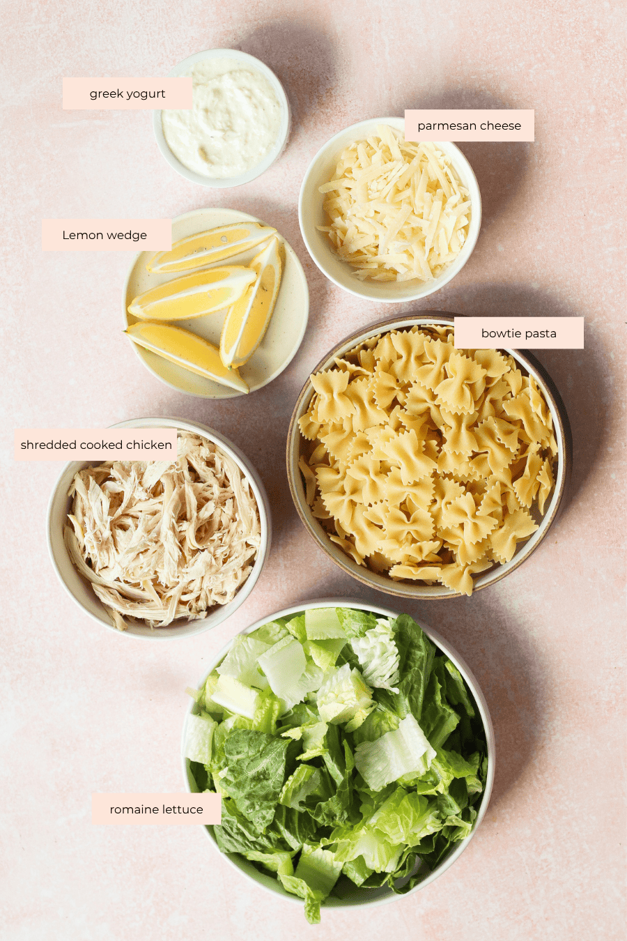 Bowls filled with Chicken Caesar pasta salad—greek yogurt, parmesan cheese, bowtie pasta, shredded cooked chicken, and chopped romaine lettuce—arranged on a light pink surface with a plate of lemon wedges. Each ingredient is labeled.