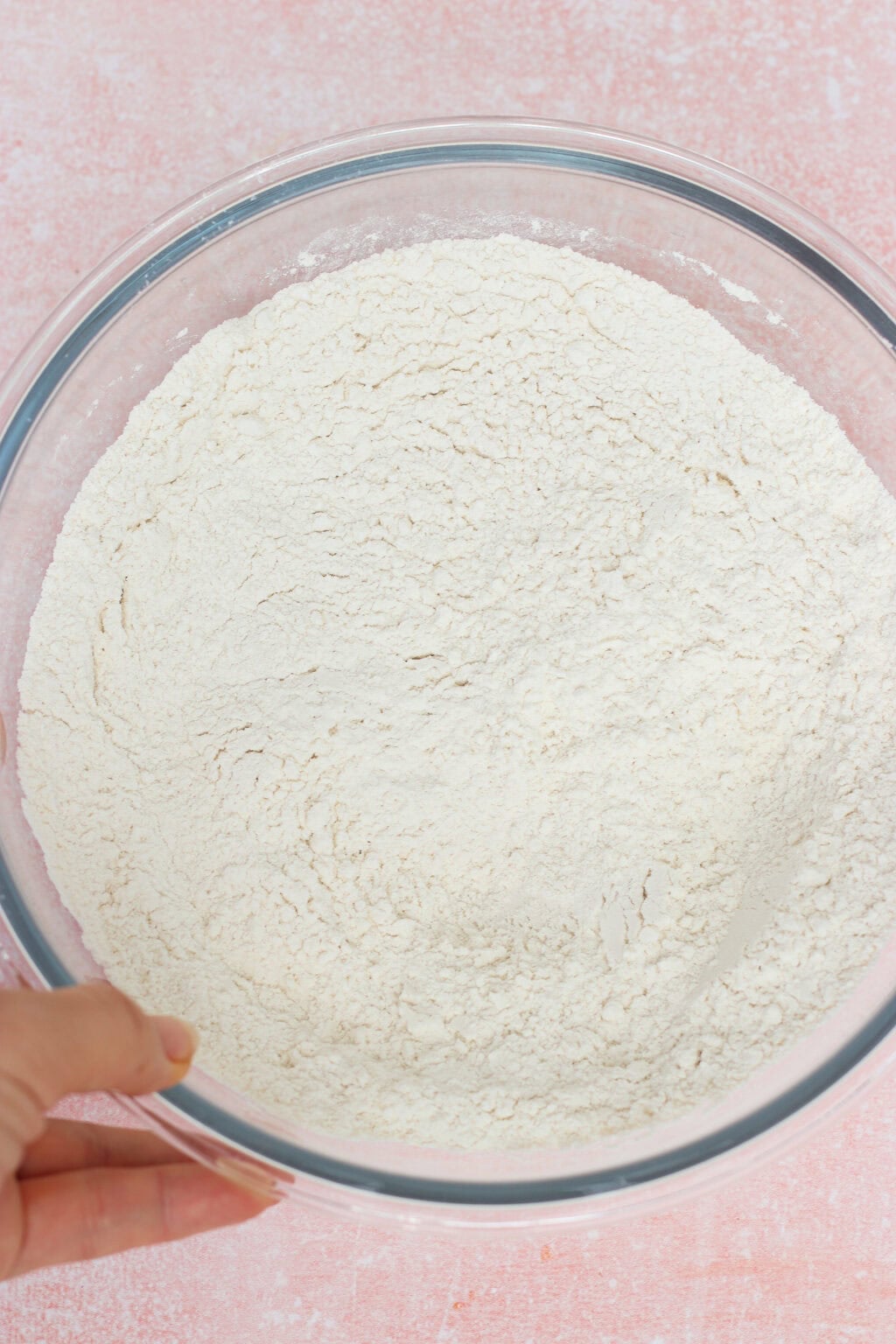 A hand holding a clear glass bowl filled with white flour, set on a light pink textured surface.