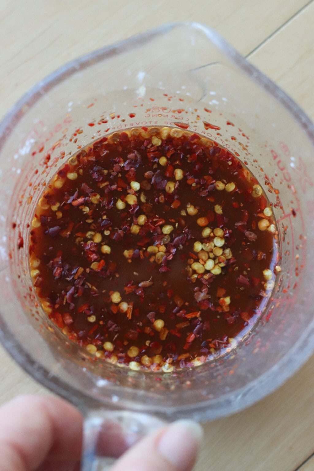 A clear measuring cup filled with a reddish-brown liquid sauce, flecked with chili flakes and seeds, is held by a hand over a light wooden surface.